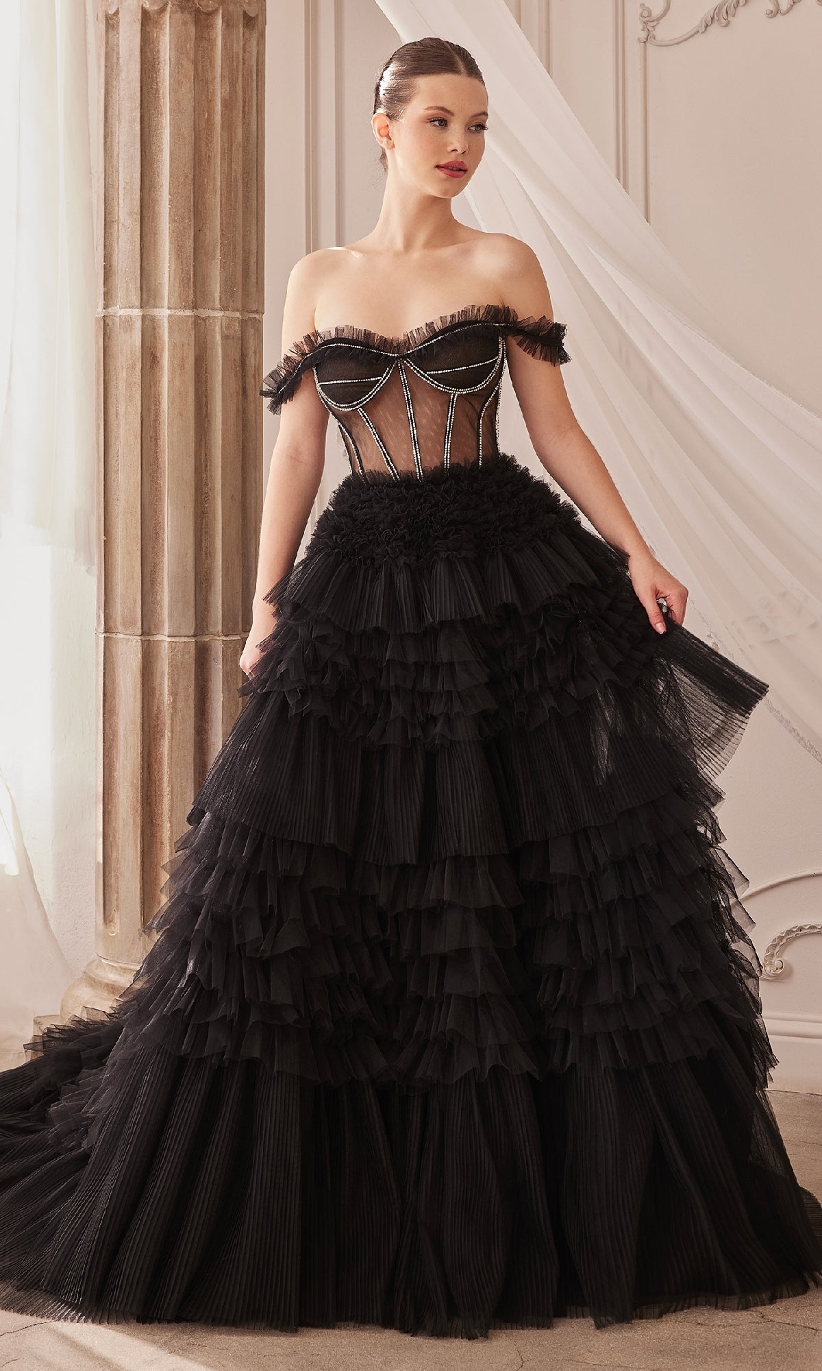 A woman stands indoors by a decorative column and draped fabric, wearing the Pastel Off-the-Shoulder Ruffled Ball Gown A1150, featuring a sheer corset bodice and multi-layered ruffled skirt.